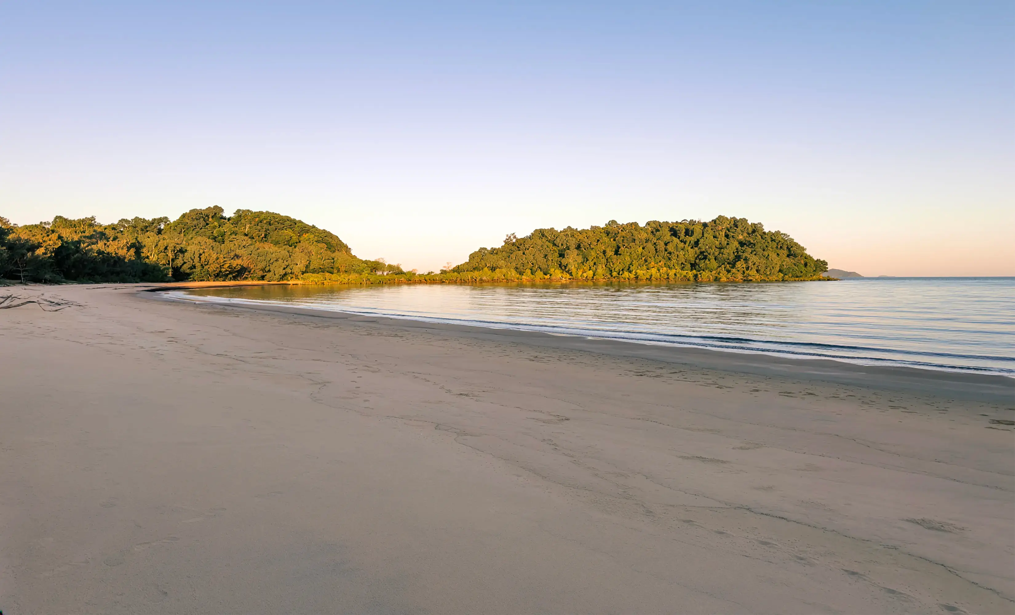 The beach at low tide, the forested headland across calm water in low light